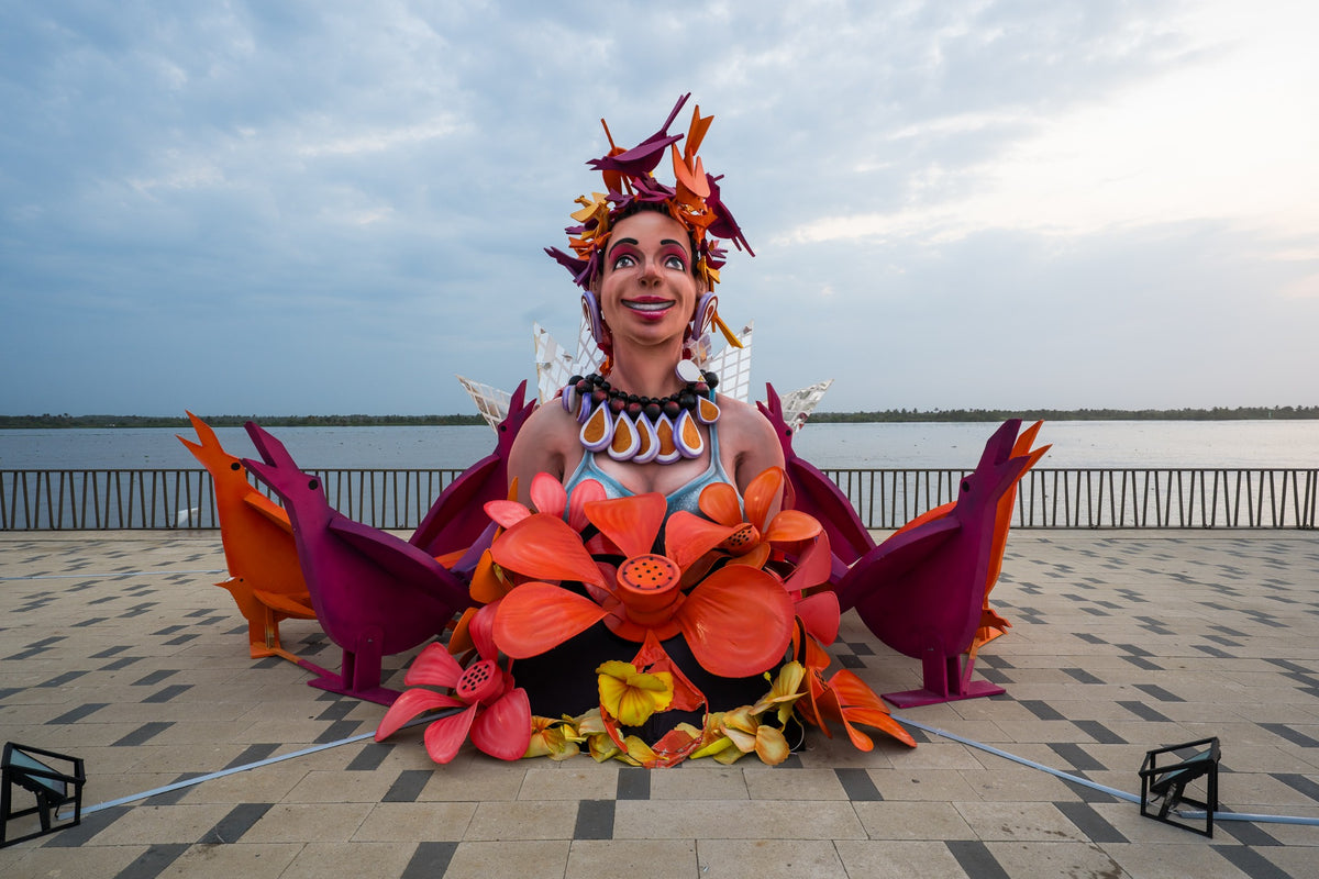 A colorful carnival sculpture with flowers and birds stands beside the river.