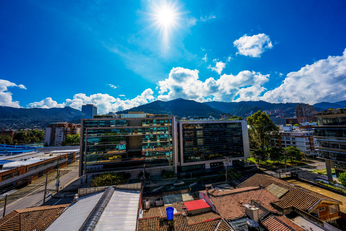 Bright sunlight above glass buildings and tiled rooftops at the foot of green mountains.