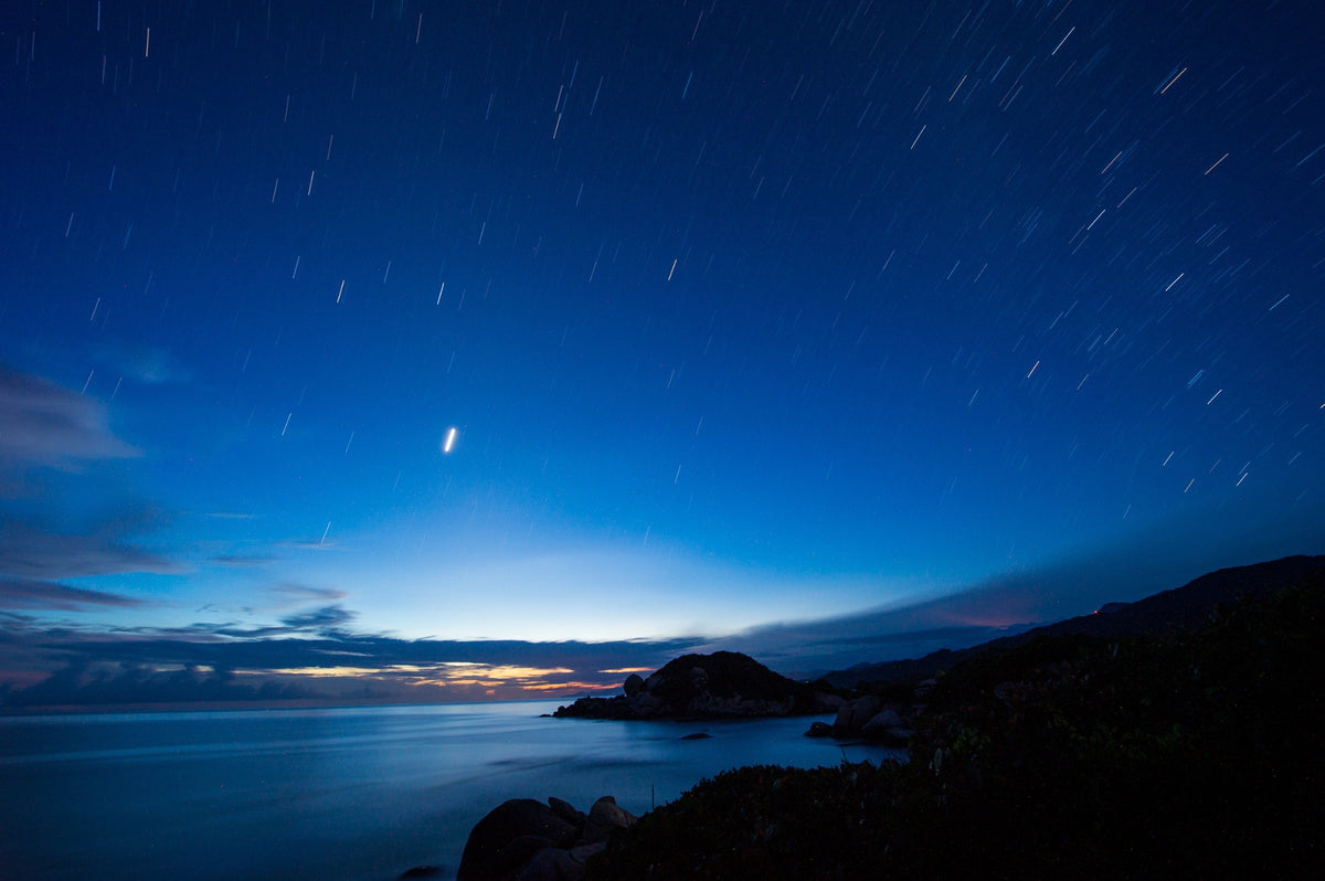 Star trails sweep above the dark coastline and long-exposure sea.