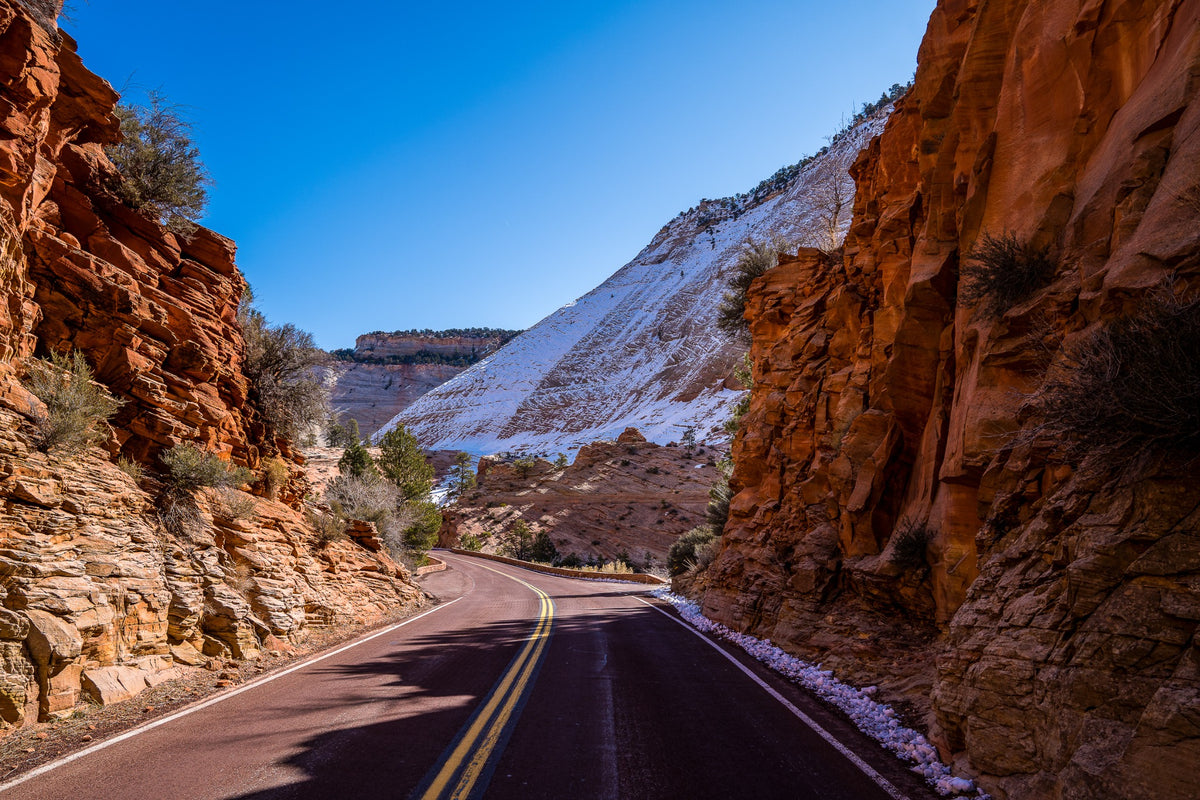 A road curves between orange rock walls and a snow-covered slope.