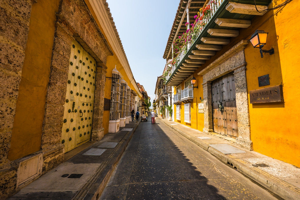 Sunlit ochre colonial street with wooden doors and flower-filled balconies.