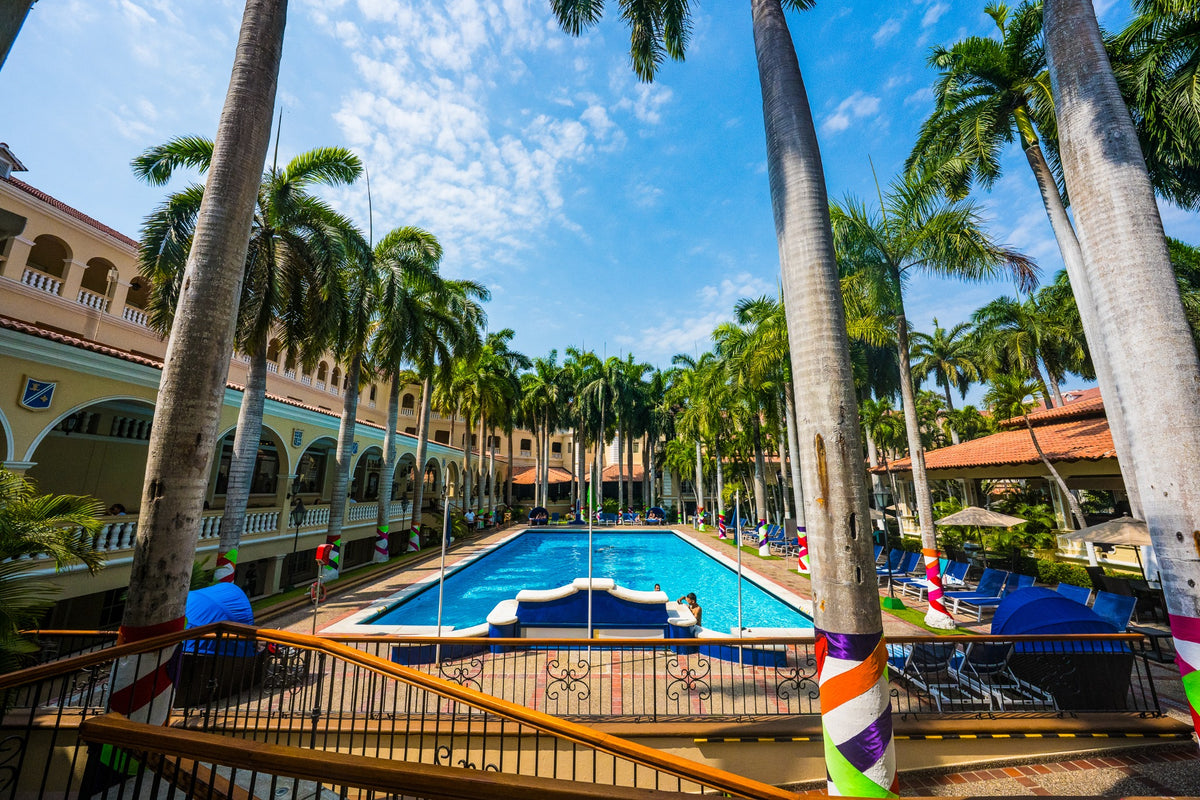 Tall palms surround a blue swimming pool and a courtyard with arched balconies.
