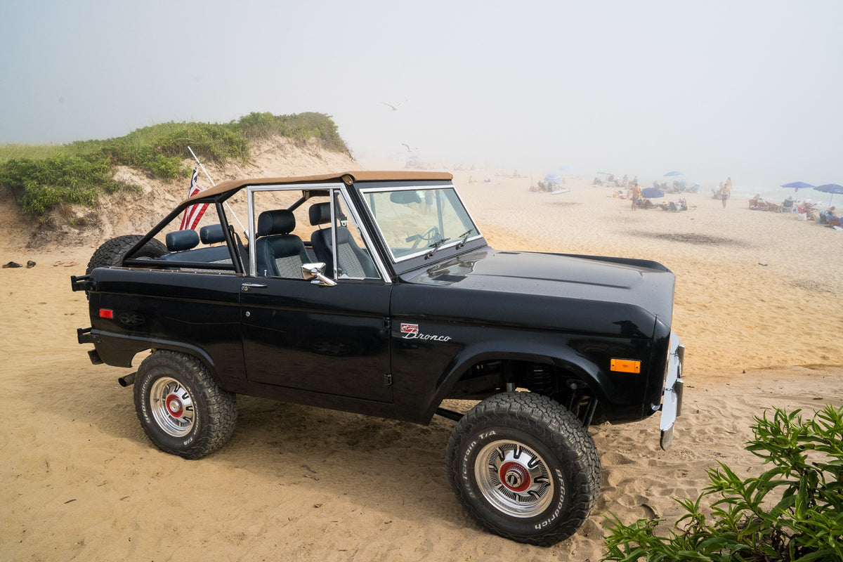 An open-top Bronco rests at the edge of the Atlantic, its dark silhouette held between sand and sea mist.