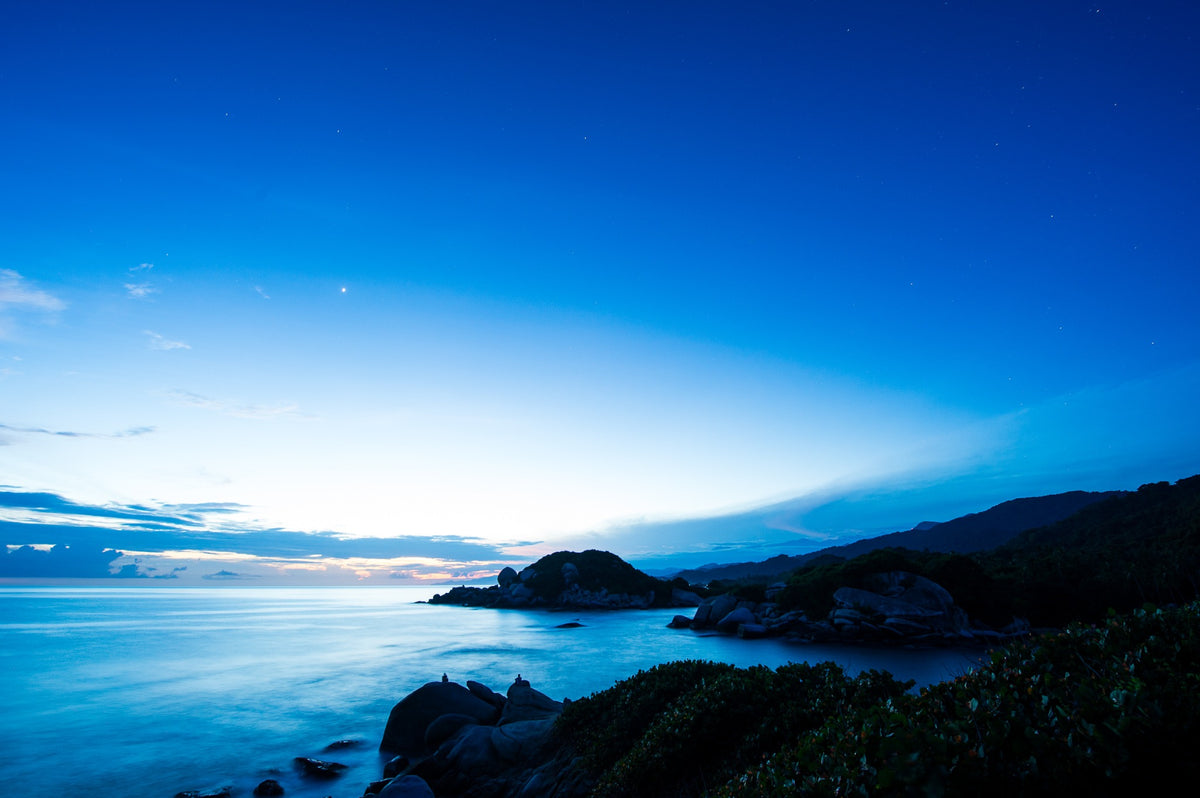 A luminous blue sky and smooth sea surround the rocky coastline.