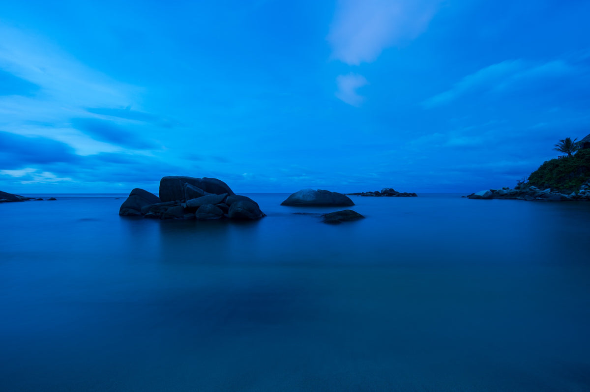 Dark offshore rocks rise from a smooth blue sea at twilight.